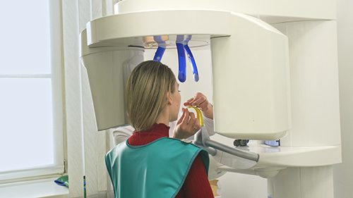 A woman in green vest eating something while standing under an x-ray machine.